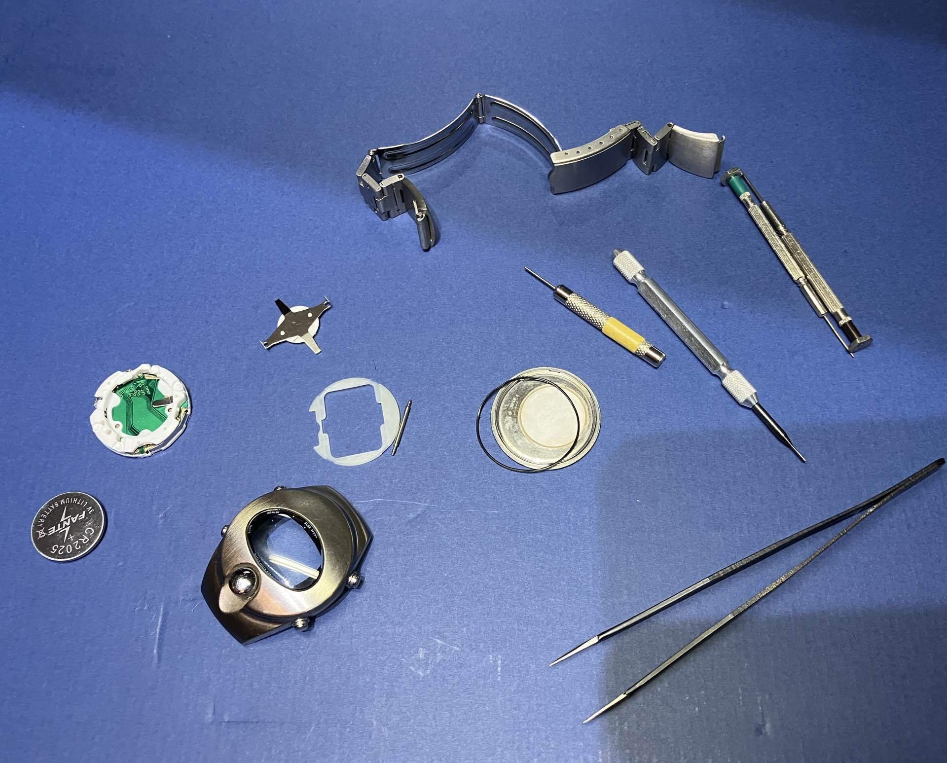 Disassembled ALBA SPOON watch with the silver circular case, removed movement, battery, band, and tools laid out on a blue work surface.