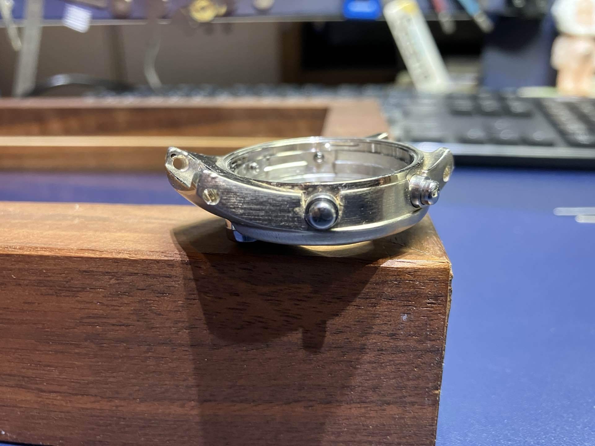Empty silver watch case resting on a wood block, with modified holes visible at the lug area.