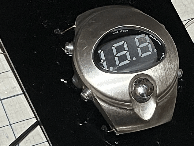 Unpowered mini voltmeter with inactive segments visible through the ALBA SPOON opening, positioned against the black mounting plate.